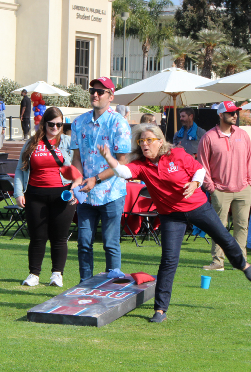 An LMU staff member standing next to a cornhole board, throwing a bean bag, with other staff members watching.